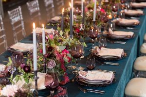 A rectangular wedding reception table featuring dark turquoise linen, a glass place setting, and a lush continuous floral centerpiece with white taper candles.